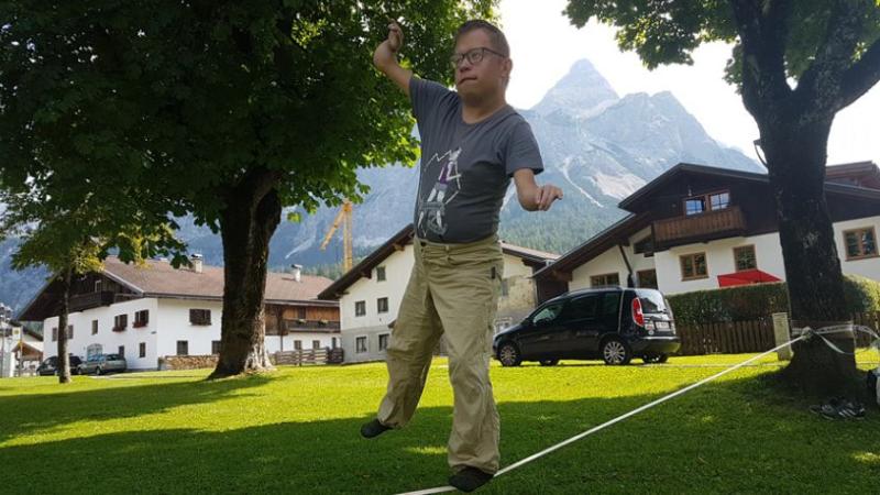 Marcel Richter auf einer Slackline, die von einem Baum aus weggeht. Marcel ist kleinwüchsig und trägt ein graues T-Shirt und eine beige Hose. Er balanciert auf der Slackline und kippt leicht nach rechts. Im Hintergrund sieht man einige Häuser und ein Bergmassiv.