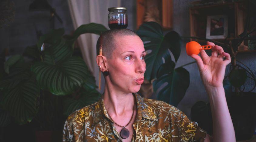 Maria hat ein Glas mit Menstruationsblut auf dem Kopf und hält ihre Menstruationstasse hoch.