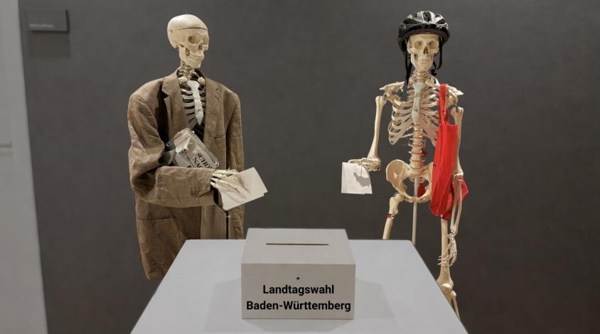 Zwei Skelette, die ihre Wahlzettel in eine Urne der Landtagswahl Baden-Württemberg schmeißen