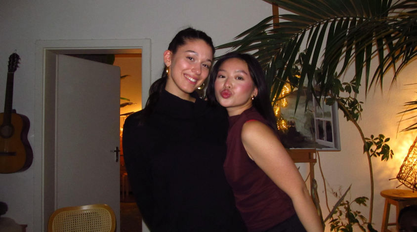 Zwei junge Frauen stehen eng nebeneinander in einem Wohnzimmer und lächeln in die Kamera. Im Hintergrund sind Pflanzen, eine offene Tür und warmes Licht zu sehen.