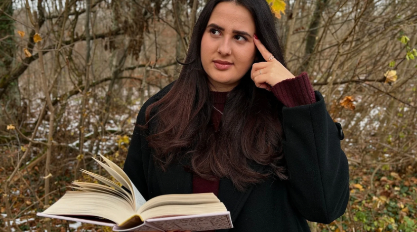 Eine Person liest ein Buch in der Natur und hat einen fragenden Gesichtsausdruck.