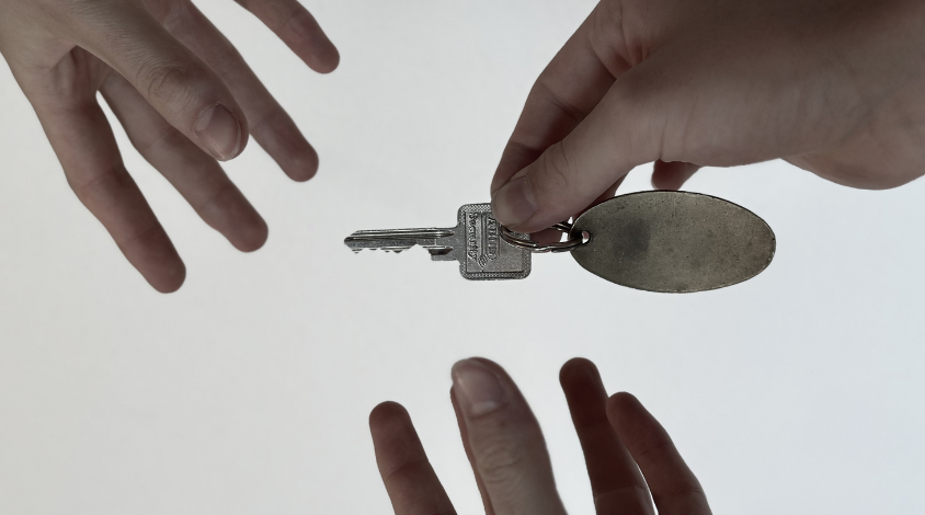 Eine Hand übergibt einen Wohnungsschlüssel mit leerem Anhänger an zwei ausgestreckte Hände … – Symbolbild für den Zugang zu Wohnraum.