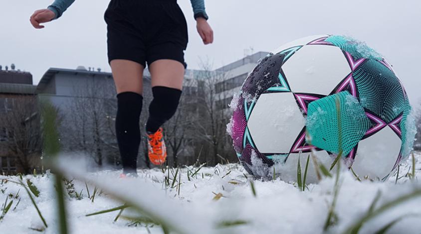 Eine Frau in schwarzen Stutzen und orangenen Fußballschuhen läuft auf einen Ball im Vordergrund des Bildes zu. Der Ball liegt auf einer schneebedeckten Wiese.