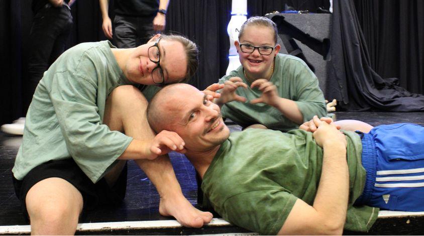 Zwei Jugendliche mit Trisomie 21 und ein Mann sitzen und liegen am Rand einer Bühne. Sie grinsen in die Kamera. Das Mädchen formt ein Herz mit ihren Händen.