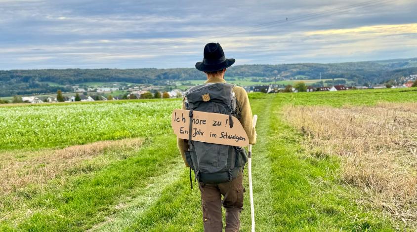Rückenansicht eines Mannes mit Wanderrucksack auf einem Feldweg. Auf seinem Rücken trägt er ein Schild mit der Aufschrift "Ich höre zu! Ein Jahr im Schweigen"