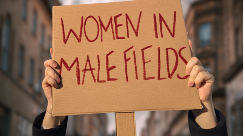 Zwei Hände halten ein Protest-Schild, auf dem der Schriftzug „WOMEN IN MALE FIELDS“ in roter Farbe und Großbuchstaben steht. Der Hintergrund zeigt verschwommene Gebäude.